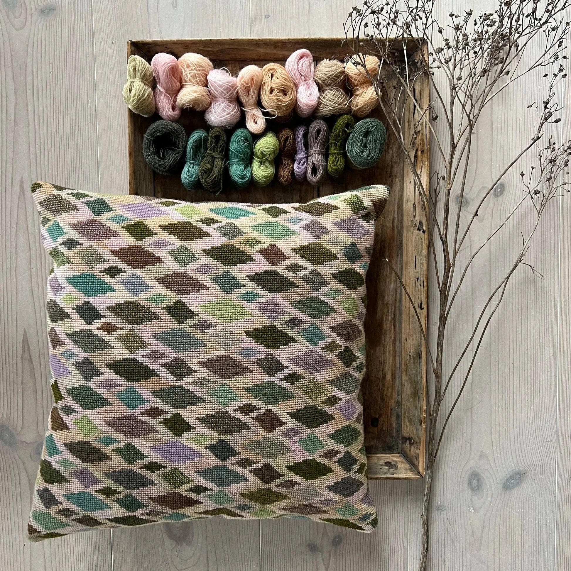Cushion no2 pute ferdig brodert med garnnøster liggende på et rustikt trefat og tregulv